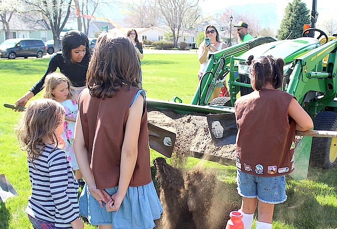 Girl Scouts shovel dirt from the bucket of a frontloader into a hole to plant a London plane tree on Arbor Day.