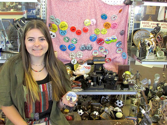 Gwen Scossa poses with her design of the 2024 Carson Valley Days button in front of some older buttons displayed at Accolades Trophies Engravings & Gifts.