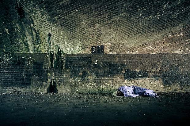 Front view wide angle image of a homeless senior adult male of caucasian ethnicity sleeping rough outdoors. He is wrapped in a blue sleeping bag on a hard floor covered with stoned, with a brick wall behind him. He has his eyes closed and has grey white hair and an unkempt white beard. Horizontal low angle image with copy space.