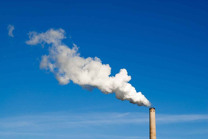 Industrial smokestack spewing white polluting smoke cloud horizontally from right to left into clear blue sky.