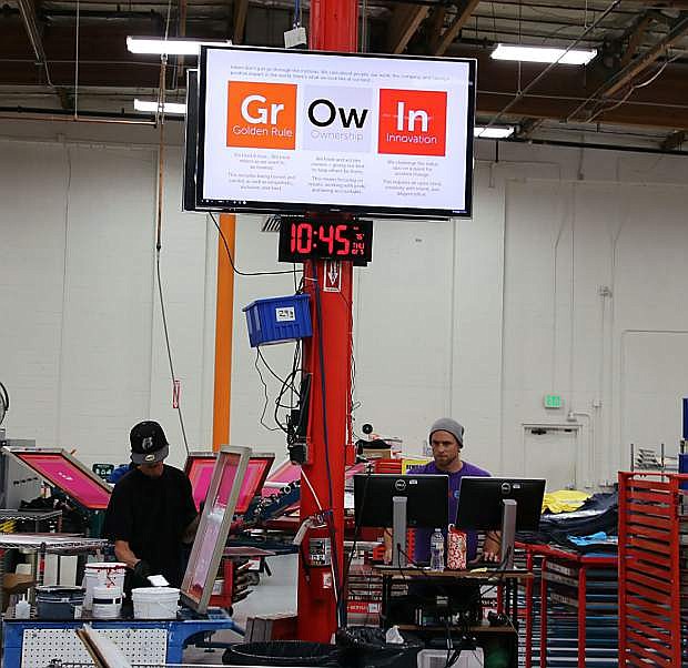 Zack Moore (left) and Jeremiah Gibson (right), two of the inkers in on the screen-printing side of CustomInk&#039;s Reno production facility, work under a sign that displays the company&#039;s core values.